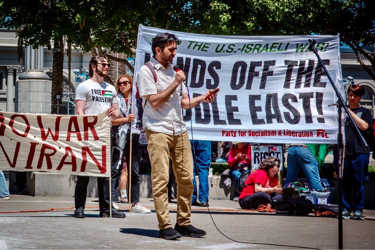 Speaker holding a microphone on a sunny day at a protest, sign in the background stays "hands off the Middle East"