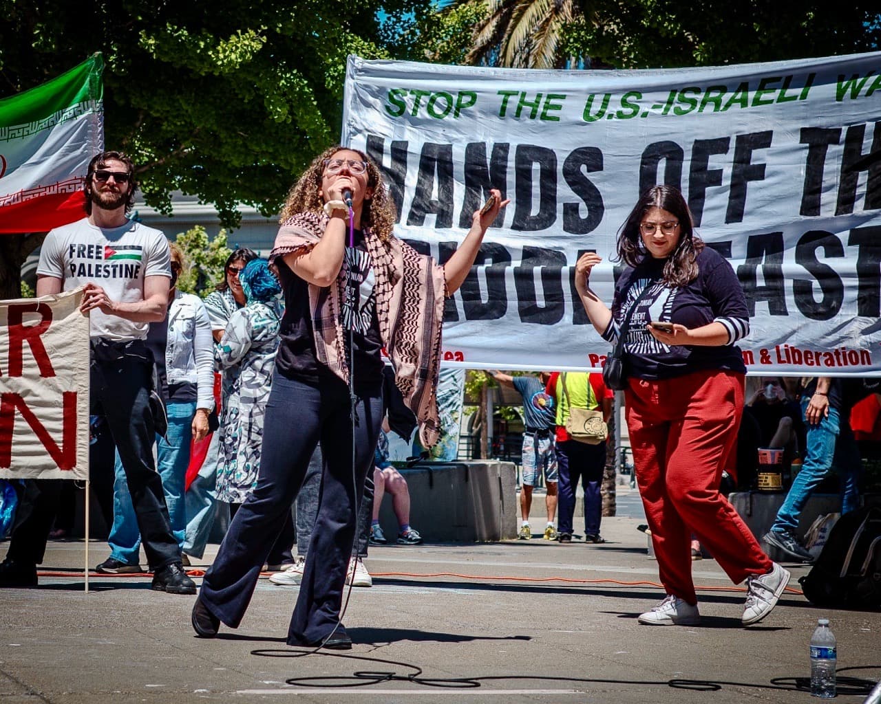 Protesters at a rally, a sign in the background says "hands off the Middle East"